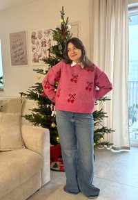 Vaaleanpunainen neule, jossa kirsikka- ja rusetti-kuvioita, yhdistetty vaaleansinisiiin leveälahkeisiin farkkuihin. Seisoo koristellun joulukuusen lähellä.