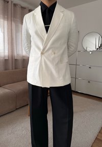 Personne portant un blazer blanc croisé, une chemise noire, une cravate noire et un pantalon noir, debout dans un salon moderne avec canapé et meuble.