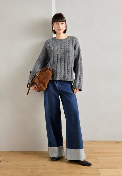 Suéter de punto grueso gris con mangas anchas, combinado con pantalones vaqueros de pierna ancha de mezclilla oscura que tienen un dobladillo de mezclilla clara. Bolso de ante marrón, zapatos negros.