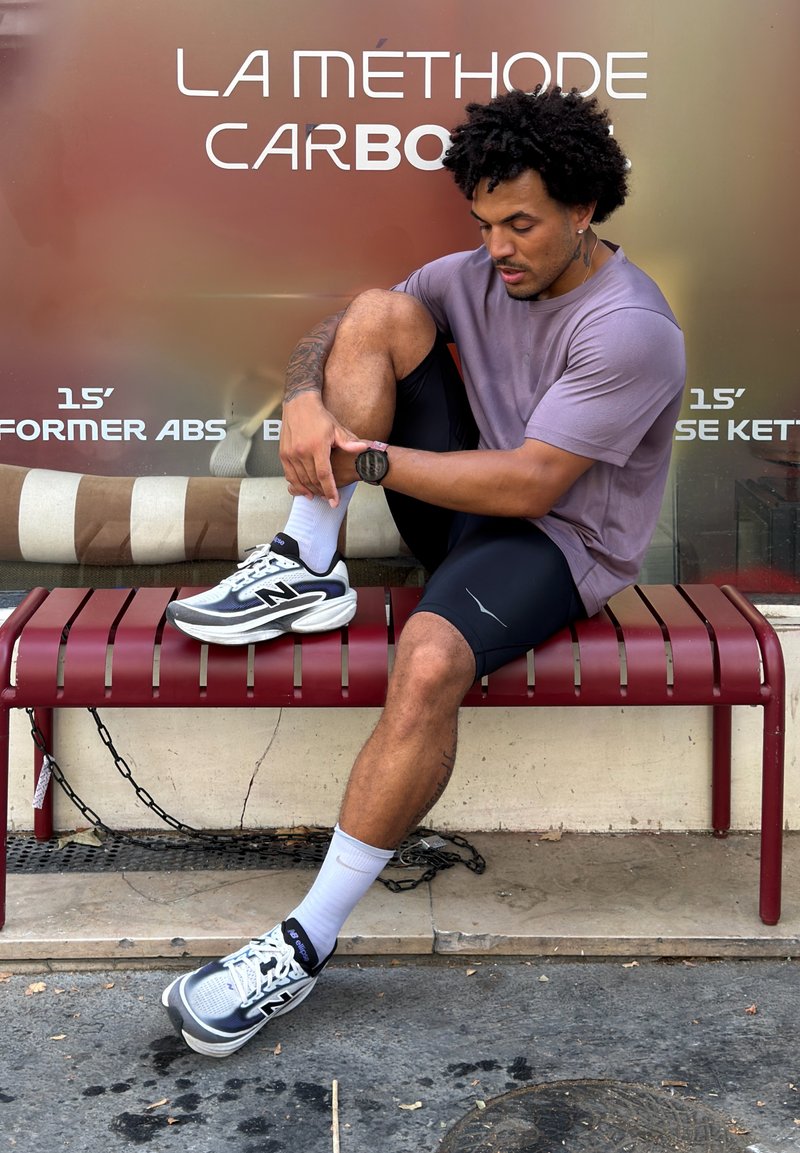 Homme assis sur un banc rouge ajustant sa chaussure, portant une tenue de sport avec des baskets, devant une publicité de fitness.