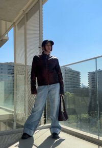 Brązowa skórzana kurtka, oversize'owe niebieskie dżinsy, czapka w kolorze czarnym i czarne buty. Trzyma brązową torbę. Szklany balkon z widokiem na miasto w tle.