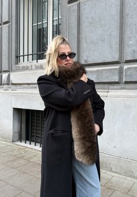 Femme blonde en manteau noir, lunettes de soleil et jeans bleus enroule une longue écharpe en fourrure marron foncé autour de son cou sur un trottoir en ville.