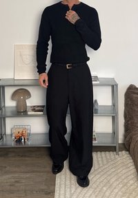 Chemise noire à manches longues côtelées avec un col rond, associée à un pantalon noir ample et une ceinture en cuir noir avec une boucle dorée.