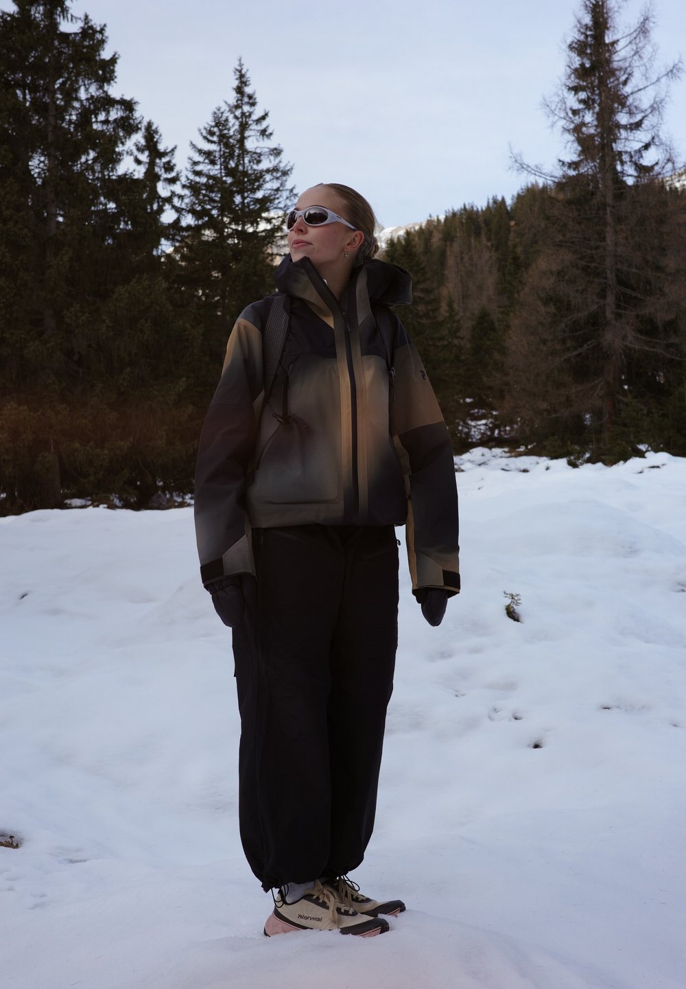 Femme portant une veste d'hiver, des gants, des lunettes de soleil et un sac à dos, debout sur la neige dans une zone montagneuse boisée, regardant vers le haut.