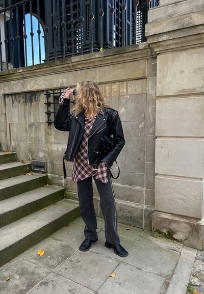 Chaqueta de motociclista de cuero negro sobre una camisa de cuadros, acompañada de jeans grises anchos con franjas laterales blancas y botas negras hasta el tobillo. Escaleras de concreto de fondo.