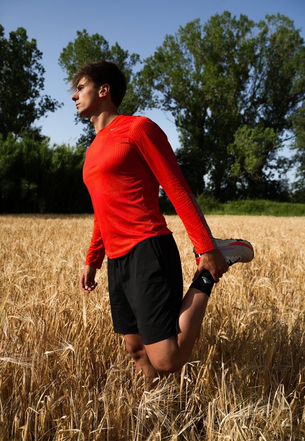 Jeune homme en chemise rouge à manches longues et short noir étire sa jambe dans un champ de blé sous un ciel bleu clair.