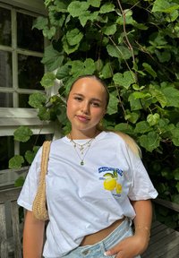 Witte katoenen t-shirt met een citroenafbeelding en blauwe tekst. Gecombineerd met lichte denim shorts en een gevlochten tas, voor een achtergrond van groene bladeren.