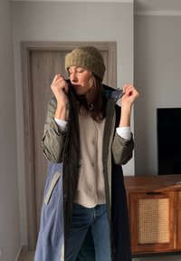 Femme portant un bonnet en tricot et plusieurs couches de vêtements d'hiver à l'intérieur, ajustant le col de son long manteau près d'une porte en bois et d'une armoire.