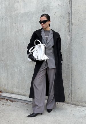 Gray wide-leg pants, gray pinstriped blazer, black long coat, black gloves, black heeled shoes, white bag, and black sunglasses against a concrete background.