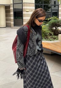 Γυναίκα που φοράει μαύρα γυαλιά ηλίου, μαύρο και άσπρο πουλόβερ με σχέδια, μαύρο graphic t-shirt, καρό φούστα και κρατά μια μεγάλη κόκκινη τσάντα σε εξωτερικό χώρο.