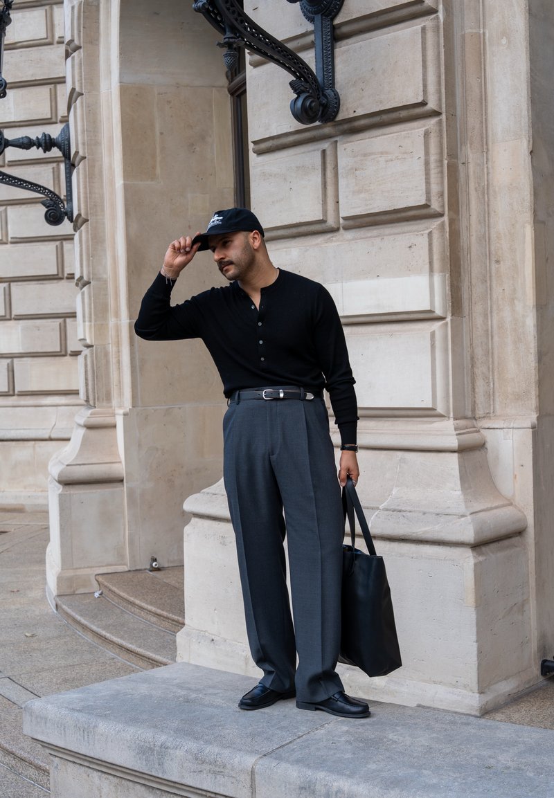 Mann in einem schwarzen Langarm-Henley-Shirt, grauen hoch geschnittenen Hosen und schwarzen Schuhen, der eine schwarze Tragetasche hält und an einer Steinmauer steht.