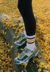 Šviesiai pilki ir žalsvai mėlyni turistiniai batai su tinklelio intarpais ir juodais akcentais, dėvimi su baltais kojinėmis, kurių dryžiai rudi ir geltoni, ant rąsto tarp geltonų lapų.