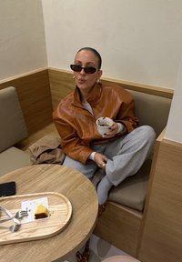 Brązowa skórzana kurtka, szare spodnie dresowe, czarne okulary przeciwsłoneczne. Trzyma kubek, siedząc na beżowej poduszce. Drewniany stół z deserem i sztućcami.