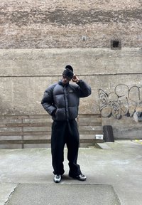 Schwarze Pufferjacke mit hohem Kragen und Logo, getragen über lockeren schwarzen Hosen. Die Accessoires umfassen eine schwarze Strickmütze und Sonnenbrille.