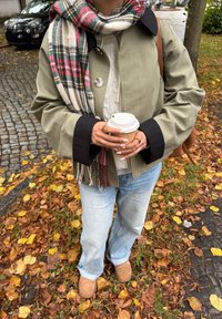 Olivenfarbener Mantel mit schwarzen Manschetten, cremefarbener Strickpullover, hellblaue Jeans und braune Wildleder-Pantoletten. Hält eine Tasse Kaffee und trägt einen karierter Schal.