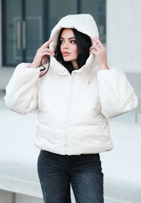 Veste à capuche blanche et gonflée avec une fermeture éclair à l'avant et des poches latérales, complétée par un tissu lisse et brillant ainsi que des manches surdimensionnées.