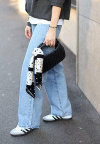Personne portant un jean bleu clair et des baskets Adidas tenant un sac à main tissé noir avec un foulard à pois blancs et noirs attaché à la poignée.
