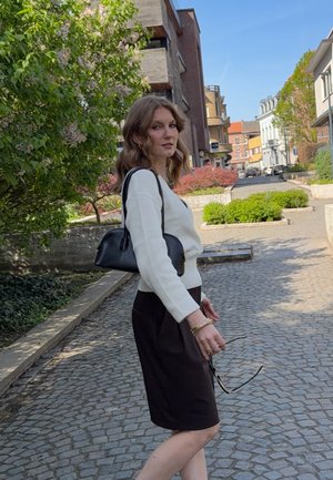 Joven mujer con cabello ondulado, que lleva un suéter blanco y una falda oscura, sosteniendo gafas de sol y un bolso negro, de pie en una calle empedrada.