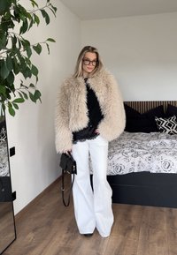 Manteau en fourrure beige duveteuse sur un haut noir, pantalon blanc à jambes larges et sac à main noir. Pièce avec un sol en bois et une literie à motifs en arrière-plan.