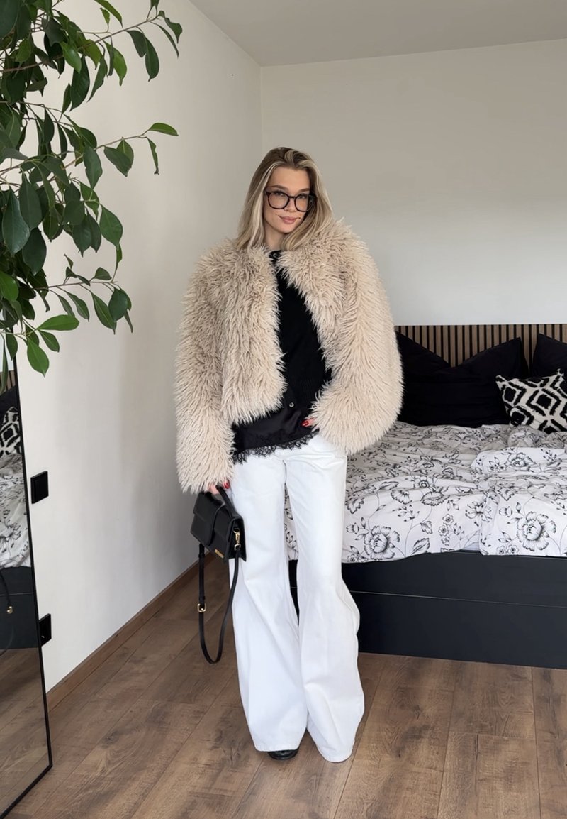 Flauschiger beiger Pelzmantel über einem schwarzen Oberteil, weiße Schlaghosen und schwarze Handtasche. Raum mit Holzboden und gemustertem Bettzeug im Hintergrund.