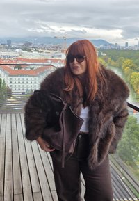 Brauner Kunstfellmantel mit weitem Kragen, getragen über einem weißen Oberteil. Dunkelbraune Hose und eine große, dunkle Handtasche. Der Hintergrund zeigt eine urbane Szenerie.