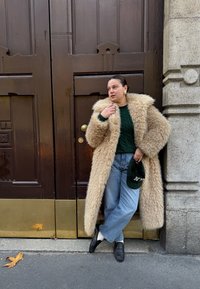 Lungo cappotto in pelliccia sintetica beige con colletto soffice, maglione lavorato a maglia verde scuro, jeans azzurri chiari e mocassini neri, con in mano un cappello verde.