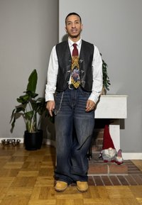 Homme portant une chemise blanche, un gilet noir, une cravate décorative, un jean large et des bottes marron clair, debout à l'intérieur près d'une cheminée avec deux décorations de gnomes.