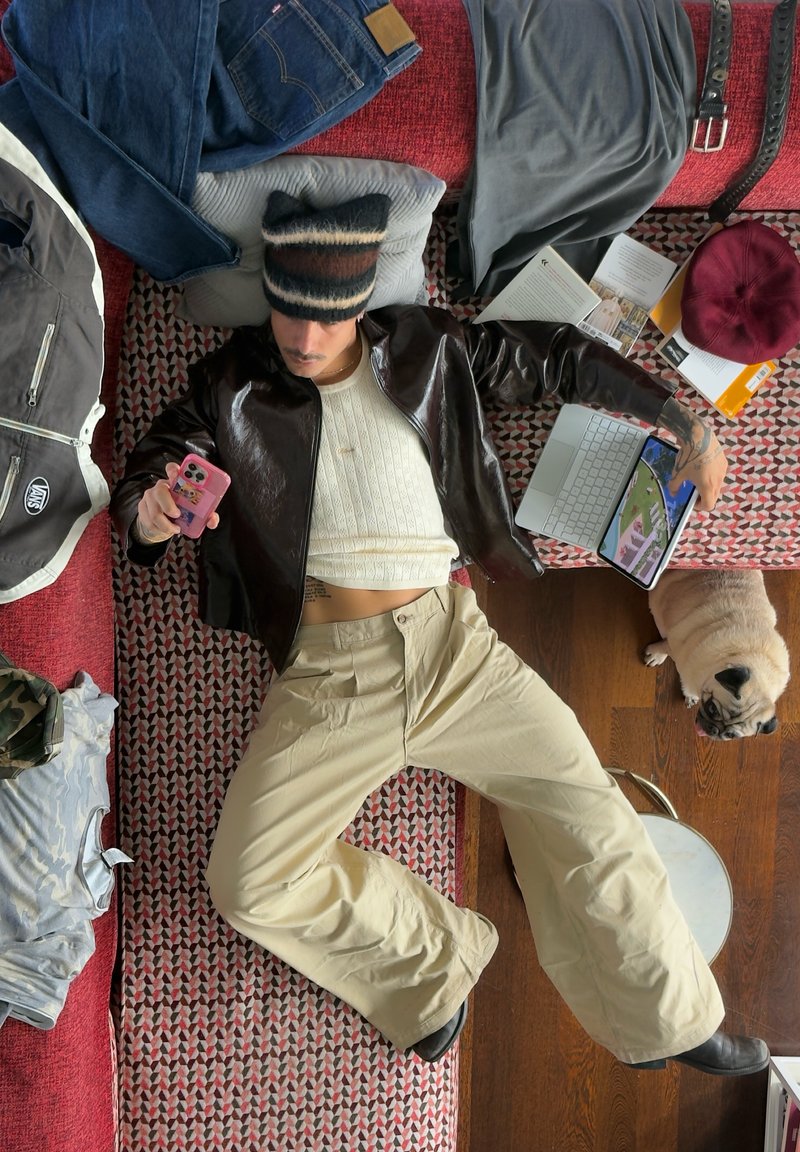 Eine Person liegt auf einem rot gemusterten Sofa, trägt eine braune Lederjacke, ein weißes, strukturiertes Oberteil und bequeme beigefarbene Hose. Um sie herum liegen Kleidung und ein Laptop.