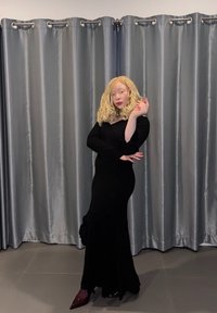 Robe noire à manches longues avec une silhouette ajustée et des poignets évasés, mise en valeur par un détail noué à la taille. Des chaussures à talons hauts complètent le look.