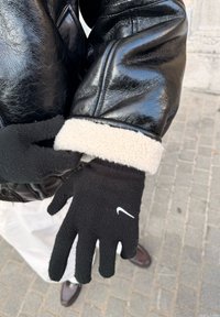 Mains portant des gants en tricot noir avec un logo blanc, ajustant une veste en cuir noire avec un revers en polaire crème sur un fond de rue pavée.
