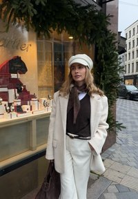 Manteau en laine beige, pull marron, pantalon blanc et casquette crème. Écharpe foncée et sac à main marron. Bijoux Cartier exposés en arrière-plan.