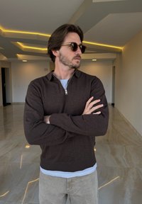 Brauner Reißverschluss-Pullover aus weichem Strickmaterial, mit lässigem Schnitt und geripptem Saum. Über einem hellblauen Hemd mit hellgrauen Hosen getragen.