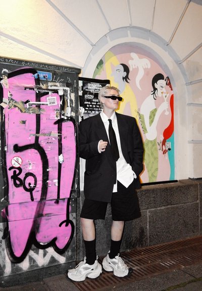 Persona que lleva un blazer negro oversized y pantalones cortos, con una camisa blanca y una corbata negra. Añadiendo un toque moderno, lleva zapatillas blancas y gafas de sol.