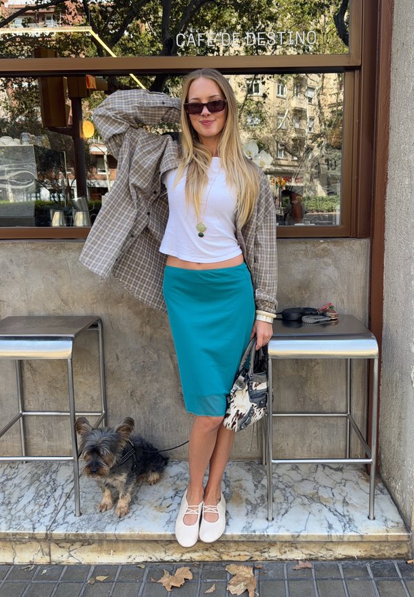 Jeune femme portant des lunettes de soleil, un haut blanc, une jupe bleu sarcelle et une chemise à carreaux, posant devant un café avec un petit chien assis à côté d'elle sur une marche en marbre.