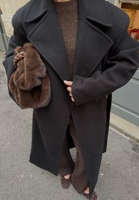 Manteau en laine noire avec un col cranté, porté sur un pull marron. Tient une pochette en fausse fourrure marron. Pantalon marron à jambes larges et chaussures à pois.
