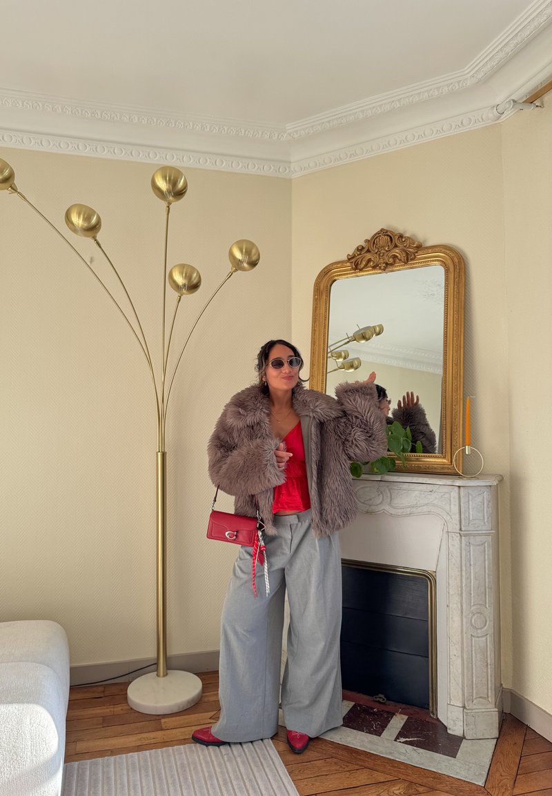 Femme portant des lunettes de soleil, un manteau en fourrure, un haut rouge, un pantalon large gris et des chaussures rouges, debout près d'une cheminée décorative avec un miroir doré et une plante.