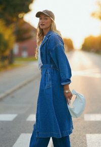 Casaco trench de ganga usado sobre uma blusa branca, com mangas longas e cintura atada. Boné cinza na cabeça, segurando uma bolsa branca, parado numa passadeira.