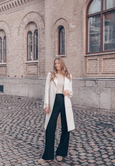 Abrigo trinchera crema sobre una blusa plisada de color beige claro y pantalones de pierna ancha negros, con una superficie de adoquines texturizada en el fondo.