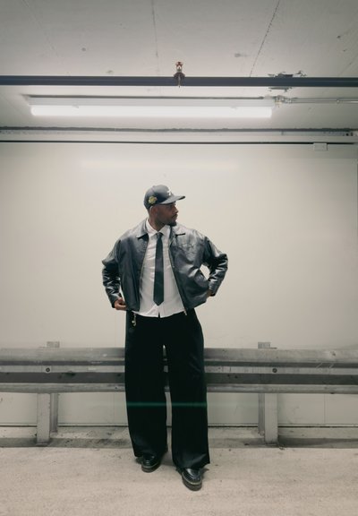 Hombre con chaqueta de cuero negra, camisa blanca, corbata negra, pantalones negros anchos y gorra negra, de pie en un aparcamiento cerca de una barrera metálica.