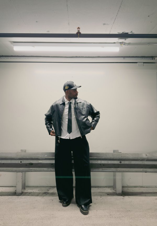 Hombre con chaqueta de cuero negra, camisa blanca, corbata negra, pantalones negros anchos y gorra negra, de pie en un aparcamiento cerca de una barrera metálica.