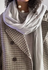 Grå, teksturert skjerf drapert over en frakk med houndstooth-mønster. Svart knapper synlige på frakken, med et lagdelt grått plagg under.