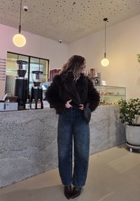 Barna műszőrme kabát, világoskék, bő szárú farmernadrág, sötét színű, magassarkú csizma. Modern kávézóbelső betonpulttal és cserepes növénnyel.