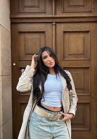 Jasnobeżowy płaszcz trencz, biały dopasowany top i jasnoniebieskie dżinsy z wysokim stanem. Czarny pasek z dużą srebrną klamrą, torebka na ramię z wzorem.