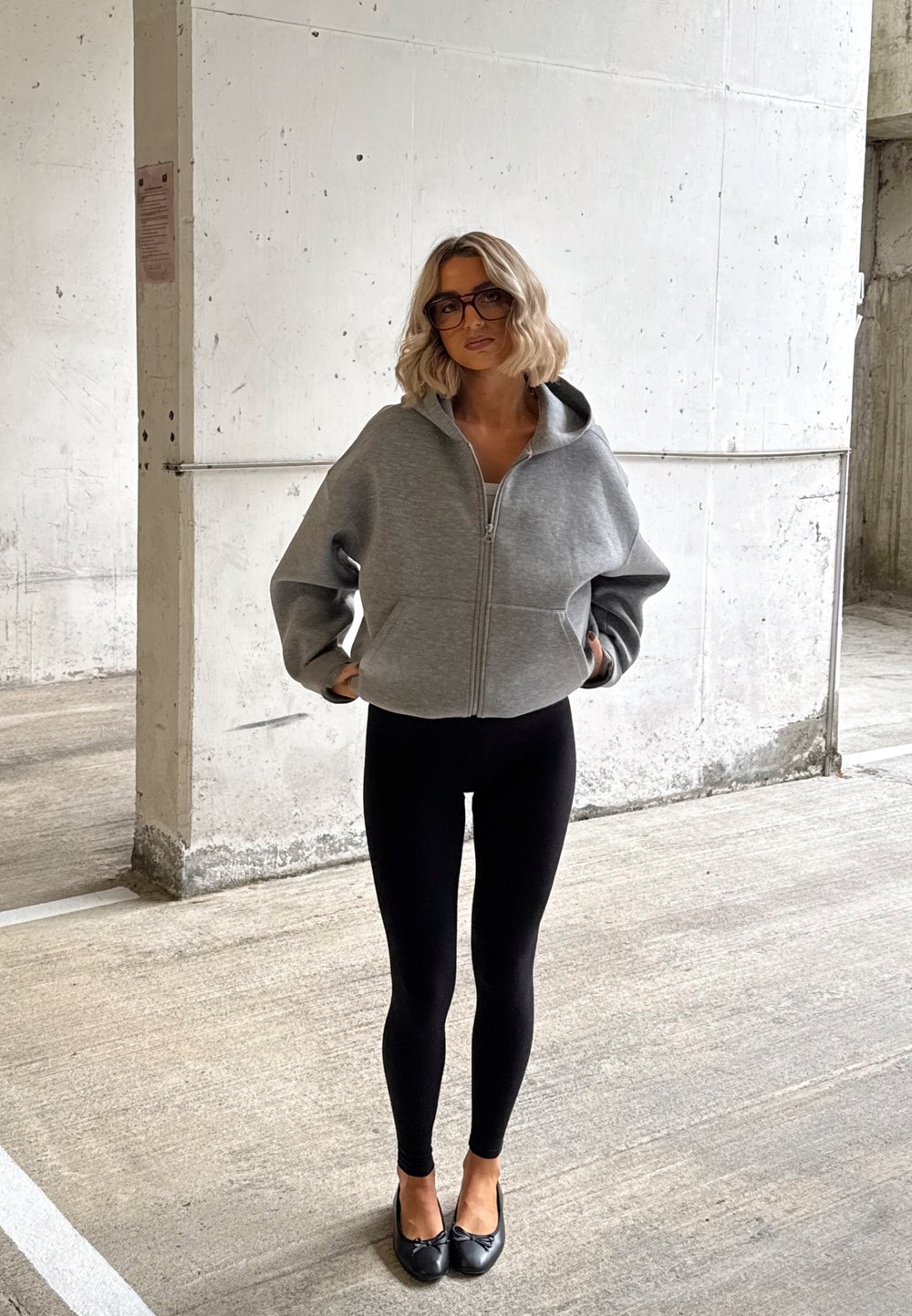 Kobieta o blond włosach ubrana w szarą, oversize'ową bluzę na zamek, czarne legginsy i czarne baleriny stoi przy betonowej ścianie na parkingu.