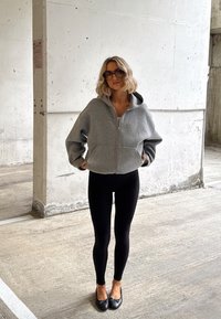 Kobieta o blond włosach ubrana w szarą, oversize'ową bluzę na zamek, czarne legginsy i czarne baleriny stoi przy betonowej ścianie na parkingu.