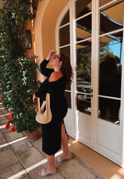 Vestido negro sin hombros con una abertura lateral, bolso de mimbre beige y sandalias deslizantes de color claro, de pie junto a una pared cubierta de hojas.