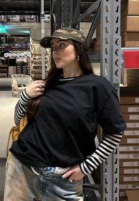 T-shirt noir surdimensionné, manches longues rayées en dessous, jean en denim et casquette de baseball camouflage ; debout contre des étagères dans un entrepôt.