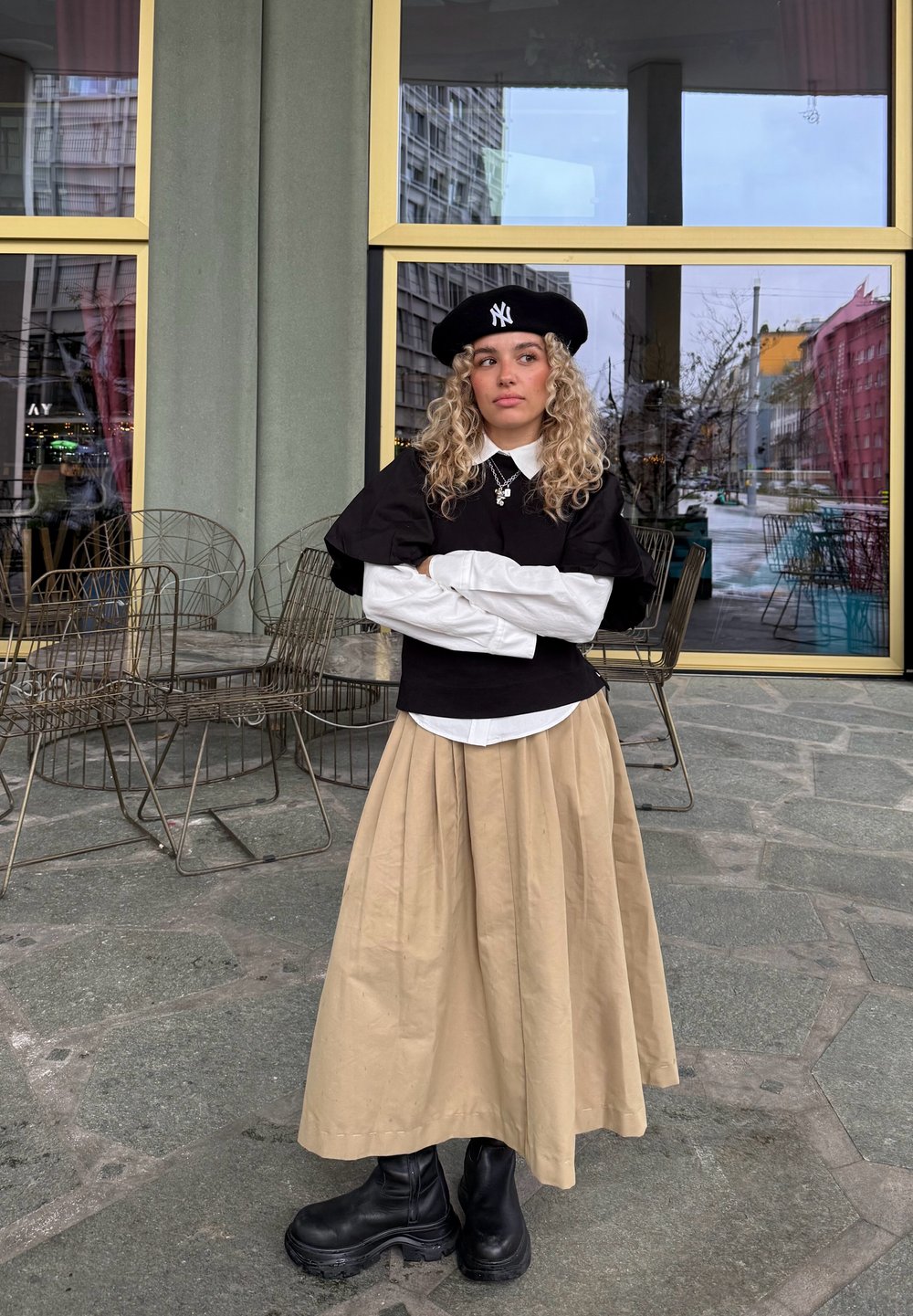 Svart genser med pufferm over en hvit skjorte med krage, beige plissett skjørt og svarte ankelstøvler. Tilbehør inkluderer en svart beret og halskjede.