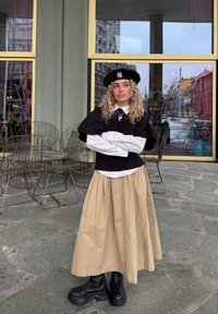 Svart genser med pufferm over en hvit skjorte med krage, beige plissett skjørt og svarte ankelstøvler. Tilbehør inkluderer en svart beret og halskjede.