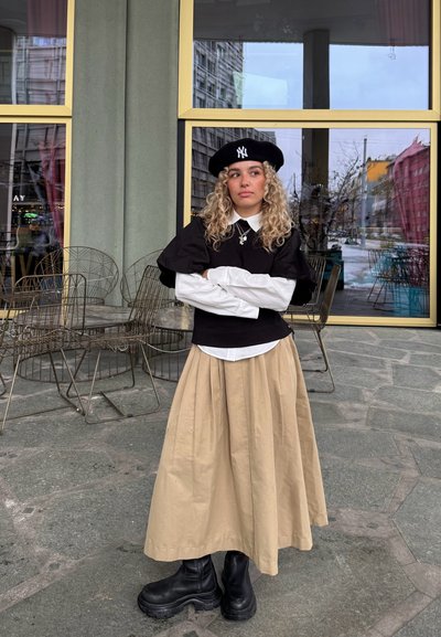 Top negro de manga abullonada sobre una camisa blanca con cuello, falda plisada beige y botines negros. Los accesorios incluyen una boina negra y un collar.